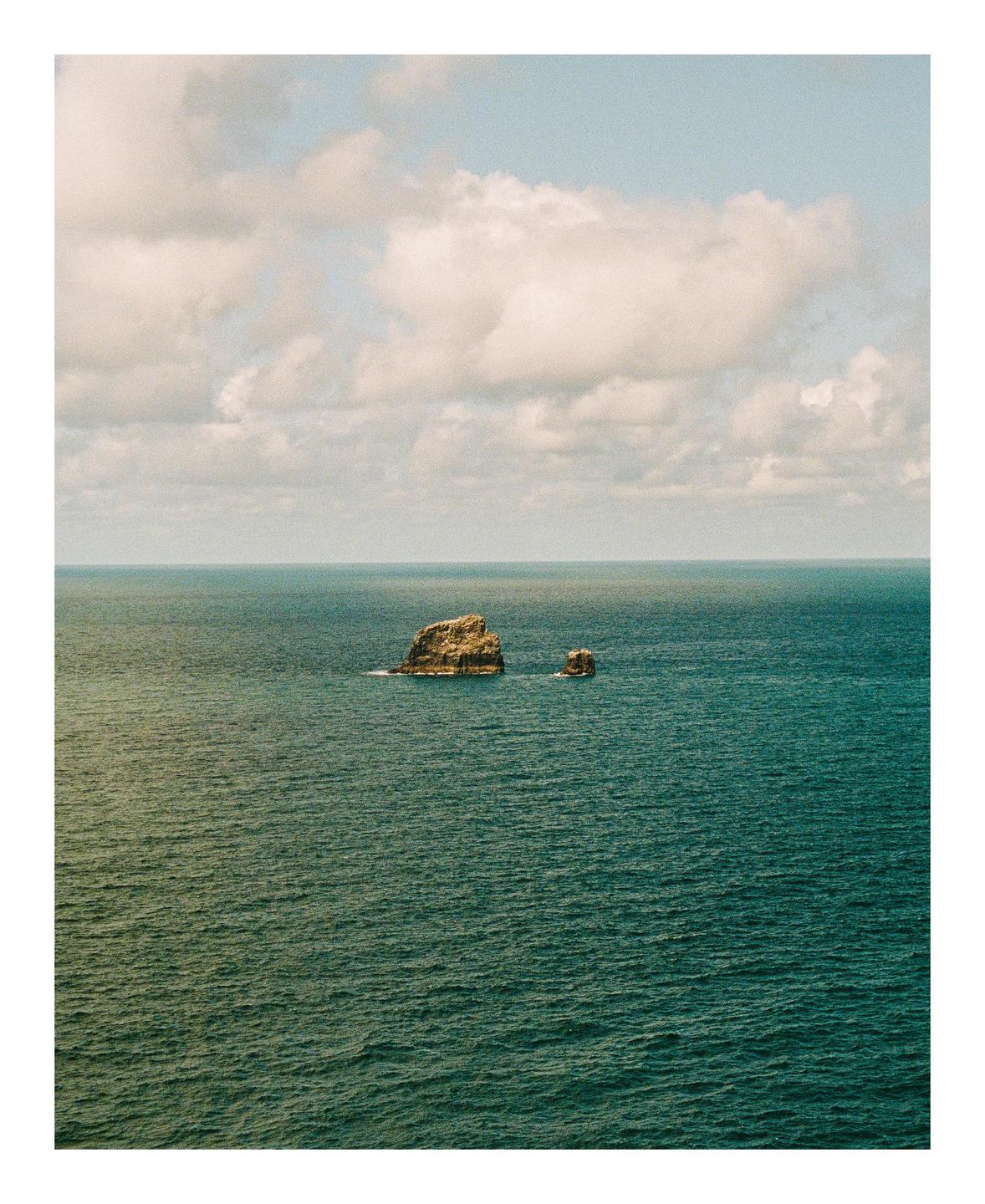 Two small rock formations emerge from the vast expanse of the ocean under a cloudy sky.