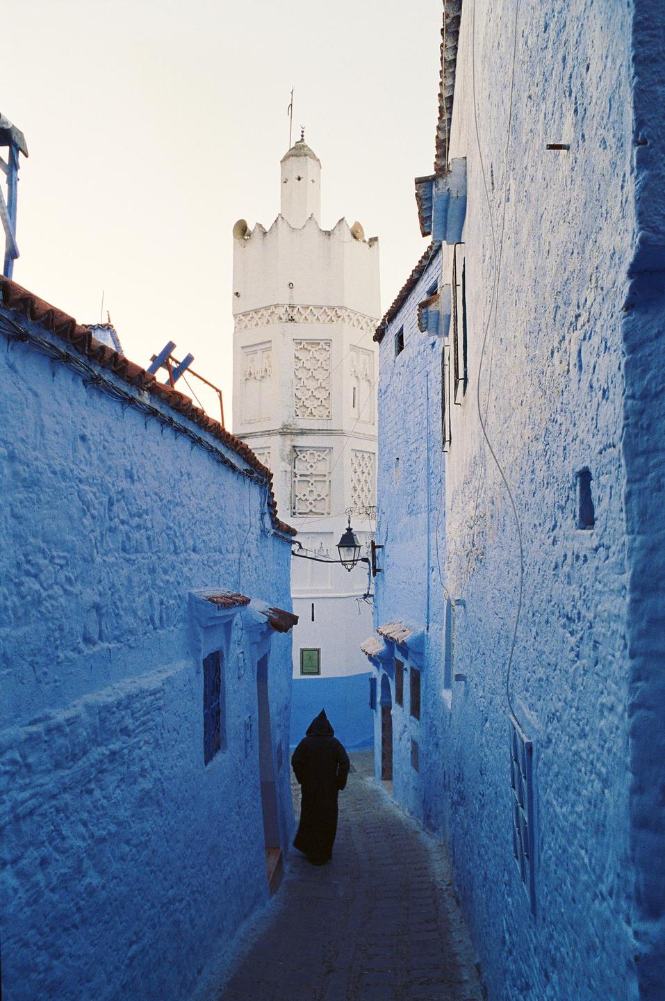 A person in a dark robe walks down a narrow blue alleyway towards a white tower.