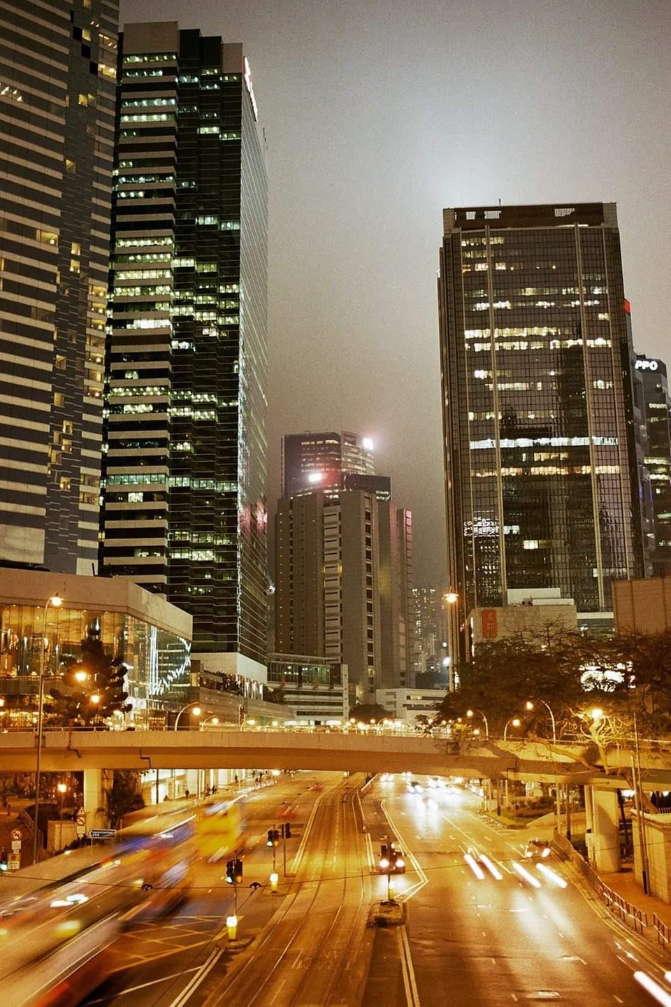 A long exposure captures light trails from vehicles on a street at night, surrounded by tall buildings in Hong Kong.