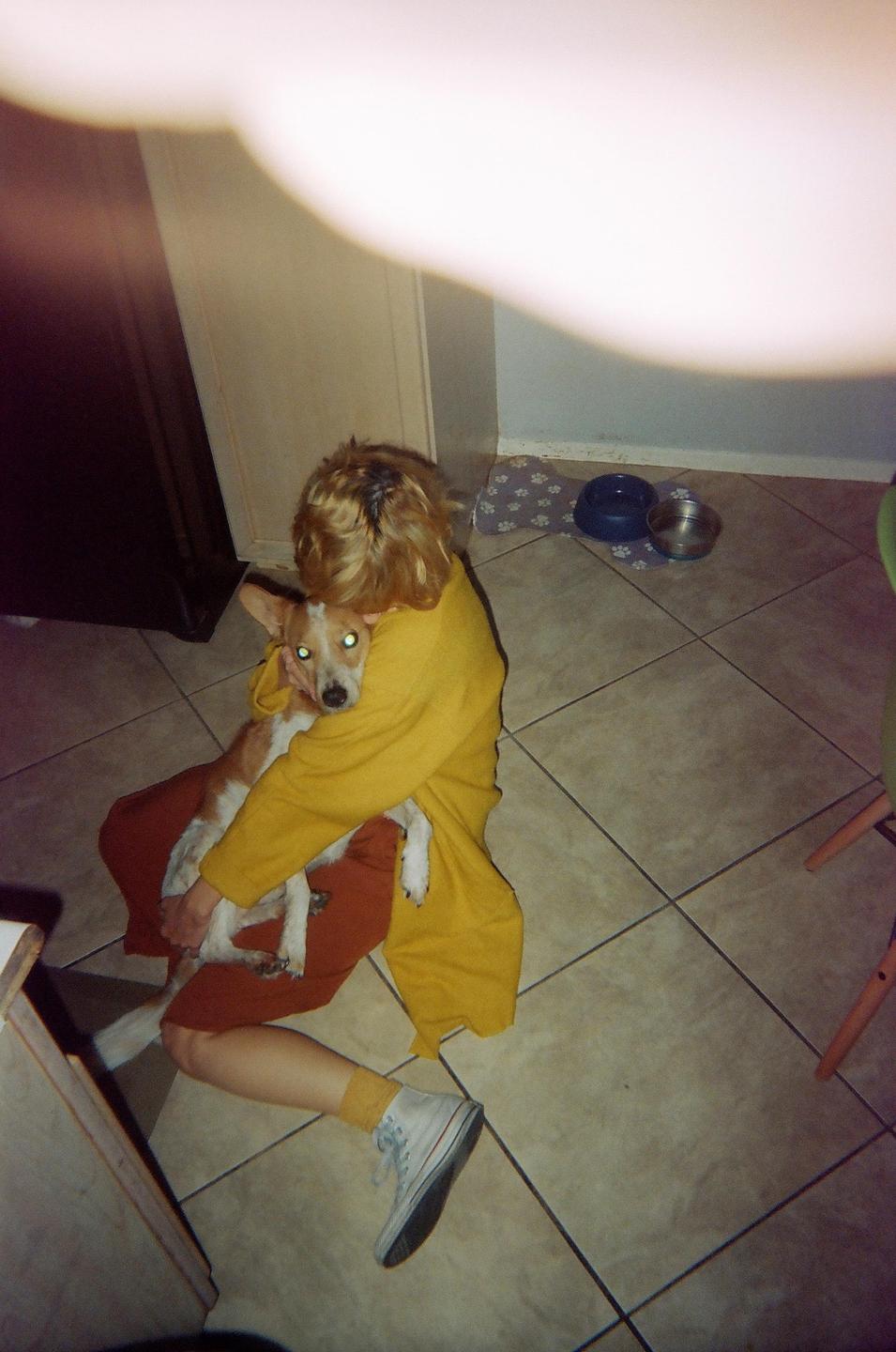 A person in a yellow top sits on a tiled floor, holding a dog with glowing eyes.