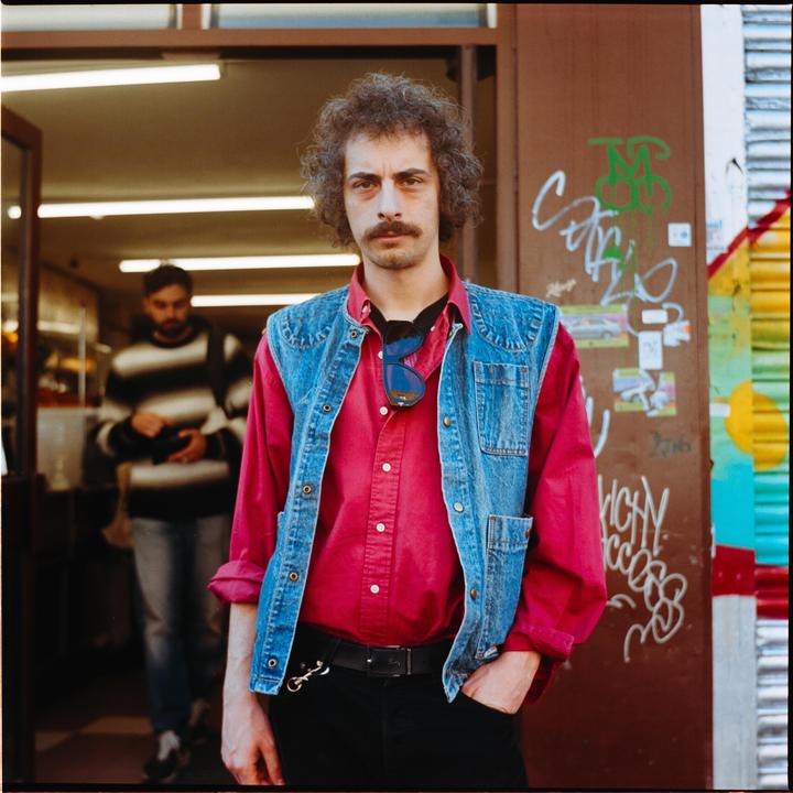 A man with curly hair and a mustache stands in front of a doorway, wearing a red shirt and a denim vest.