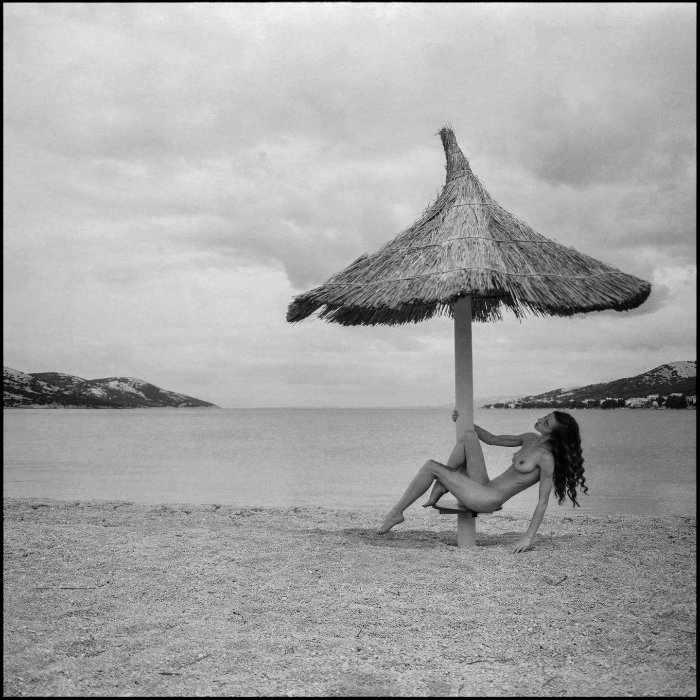 A nude woman with long hair poses under a straw umbrella on a beach with mountains in the background.