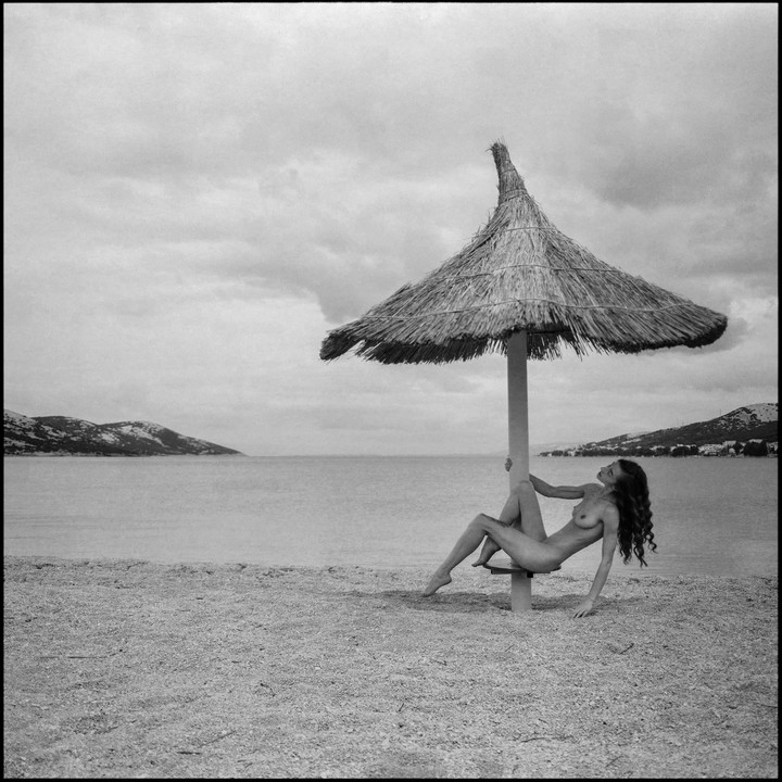 A nude woman with long hair poses under a straw umbrella on a beach with mountains in the background.