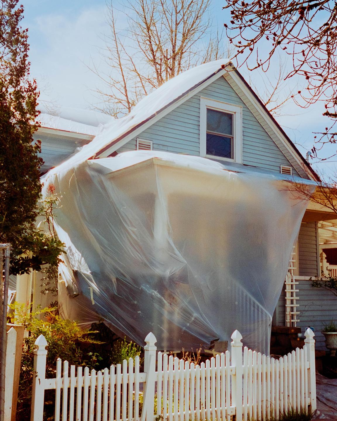 A light blue house is partially covered in clear plastic sheeting, with a white picket fence in the foreground.