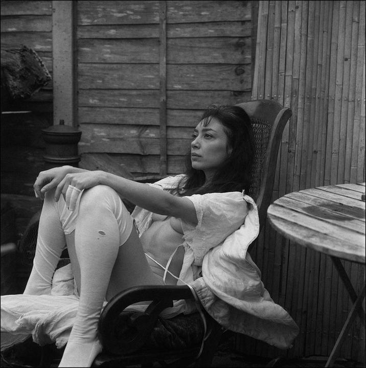 A woman sits in a chair with a white shirt draped over her shoulders, next to a small wooden table.