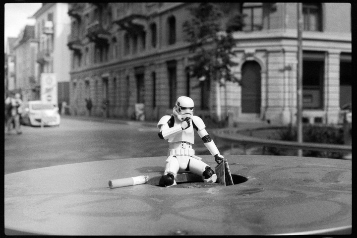 A stormtrooper action figure is kneeling on a manhole cover next to a cigarette on a city street.
