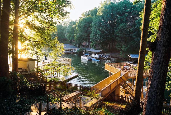 The sun shines through trees onto a lake with docks and boats, with a couple sitting on a deck in the foreground.