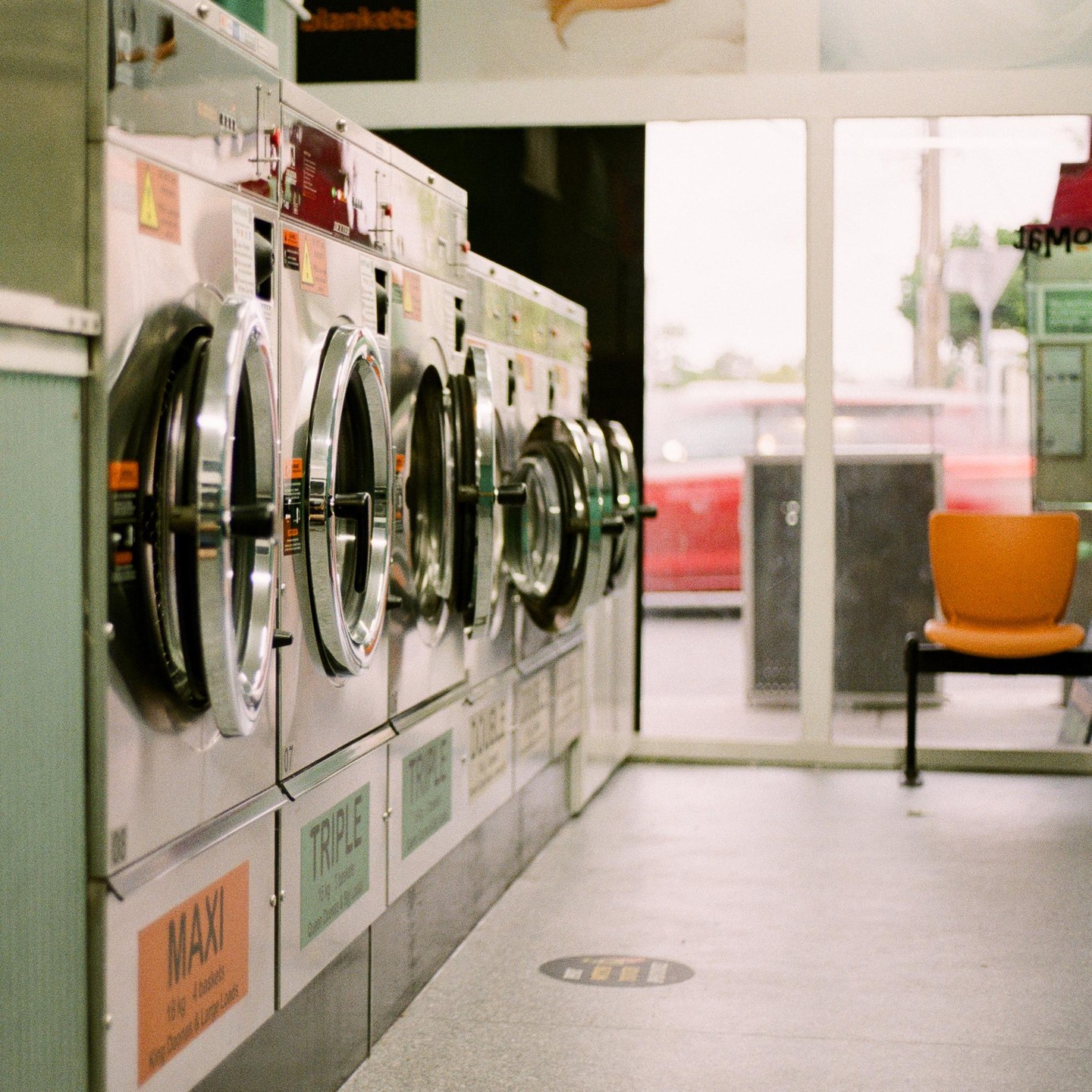 A row of washing machines stands in a laundromat with an orange chair visible through a window.
