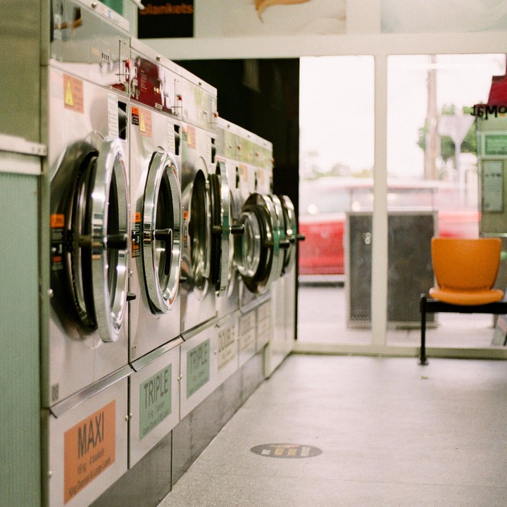 A row of washing machines stands in a laundromat with an orange chair visible through a window.