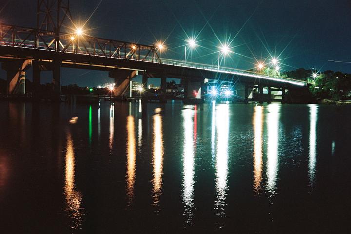 A bridge with car light trails crosses over water at night, with reflections of the lights visible on the water.