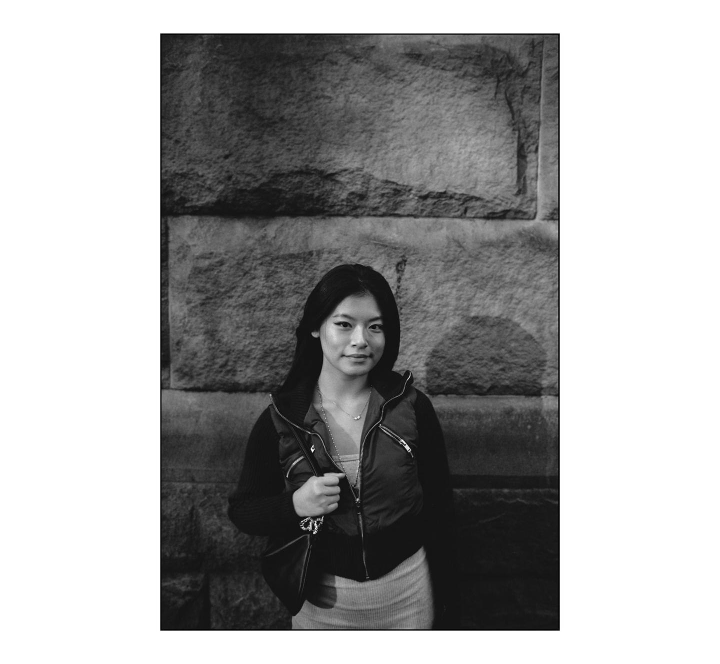 A woman stands in front of a stone wall, looking at the viewer.