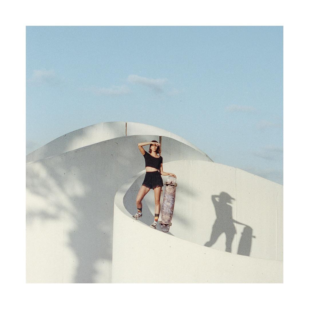 A woman stands on a white curved structure, holding a skateboard and shielding her eyes from the sun.