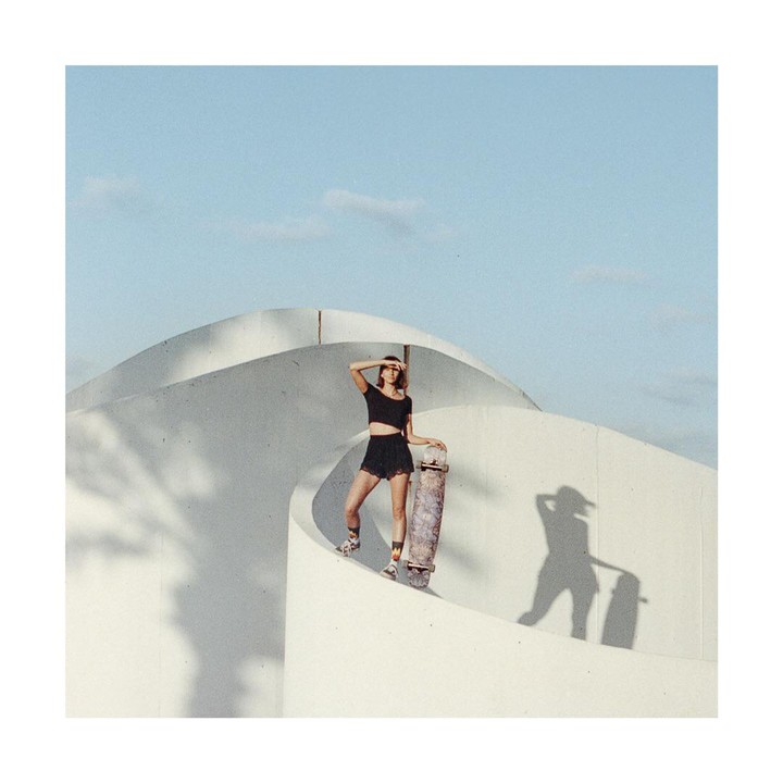 A woman stands on a white curved structure, holding a skateboard and shielding her eyes from the sun.