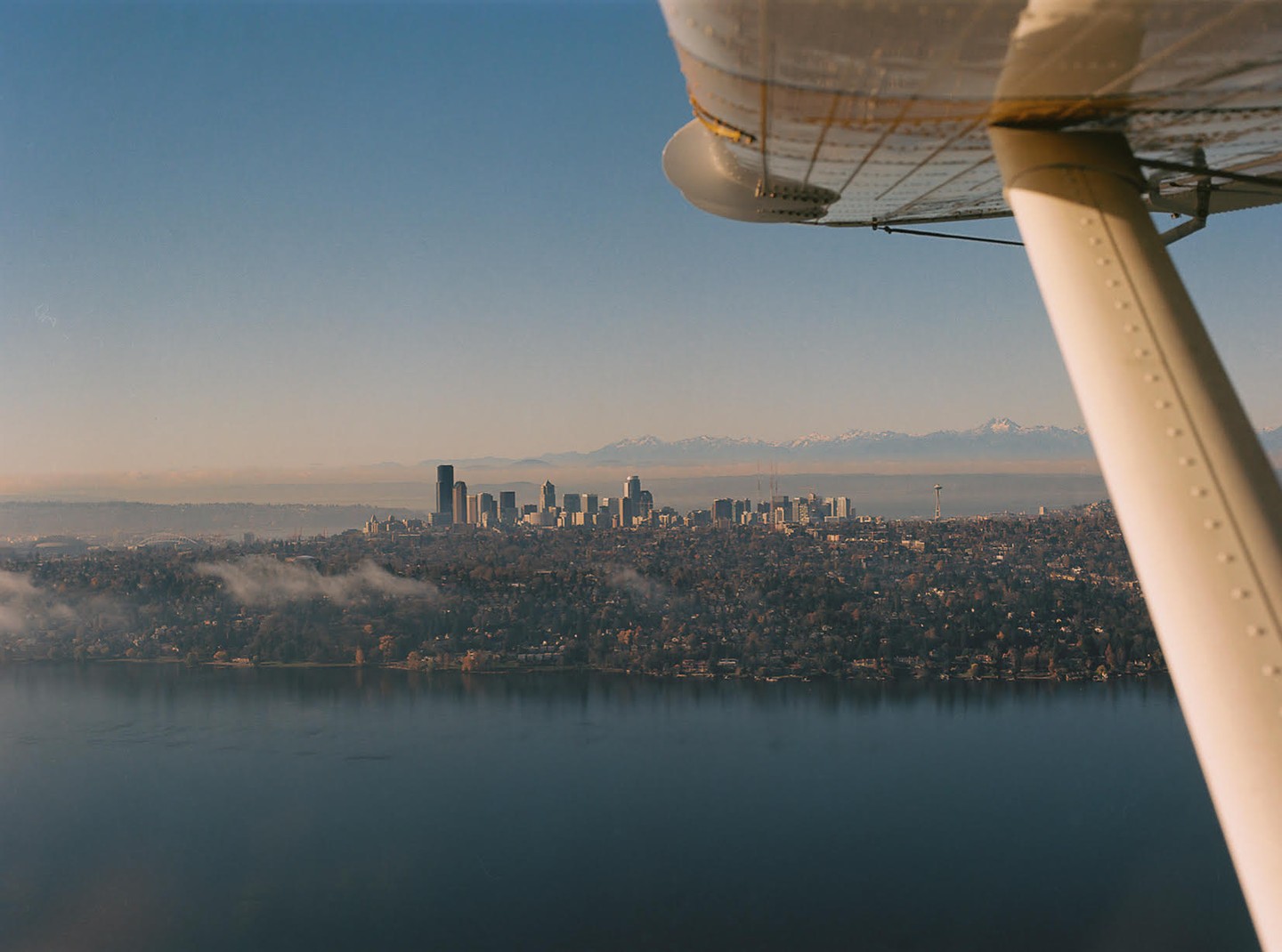 An airplane wing is visible in the foreground of a cityscape with a body of water and mountains in the background.