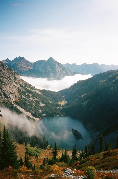 A mountain lake is surrounded by pine trees and autumn foliage, with fog rolling through the valley.
