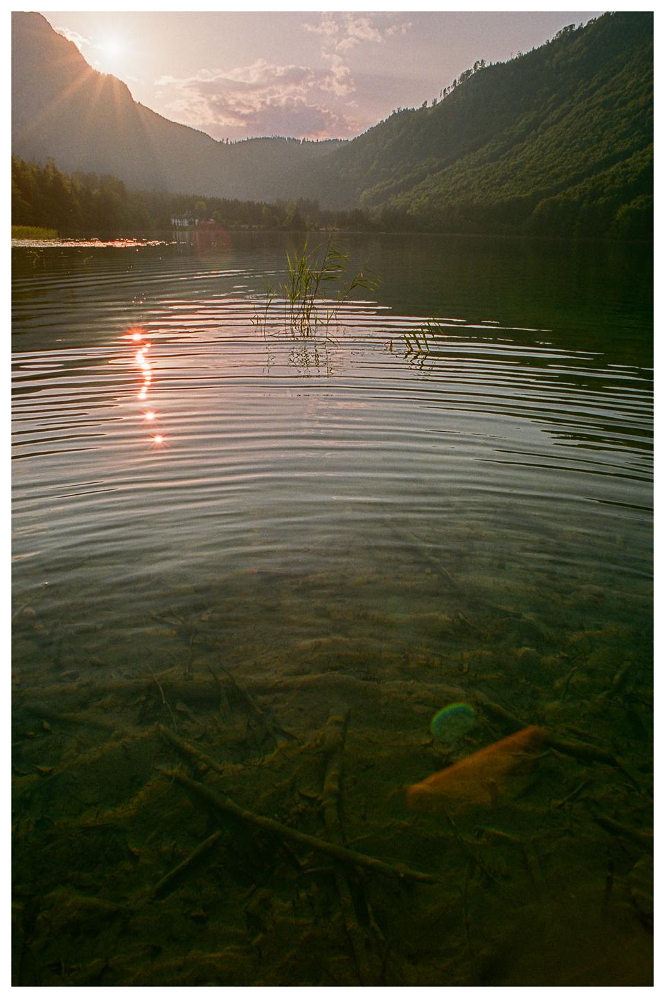 The sun sets over a lake surrounded by mountains, with the sun's reflection visible on the water's surface and submerged branches visible below.