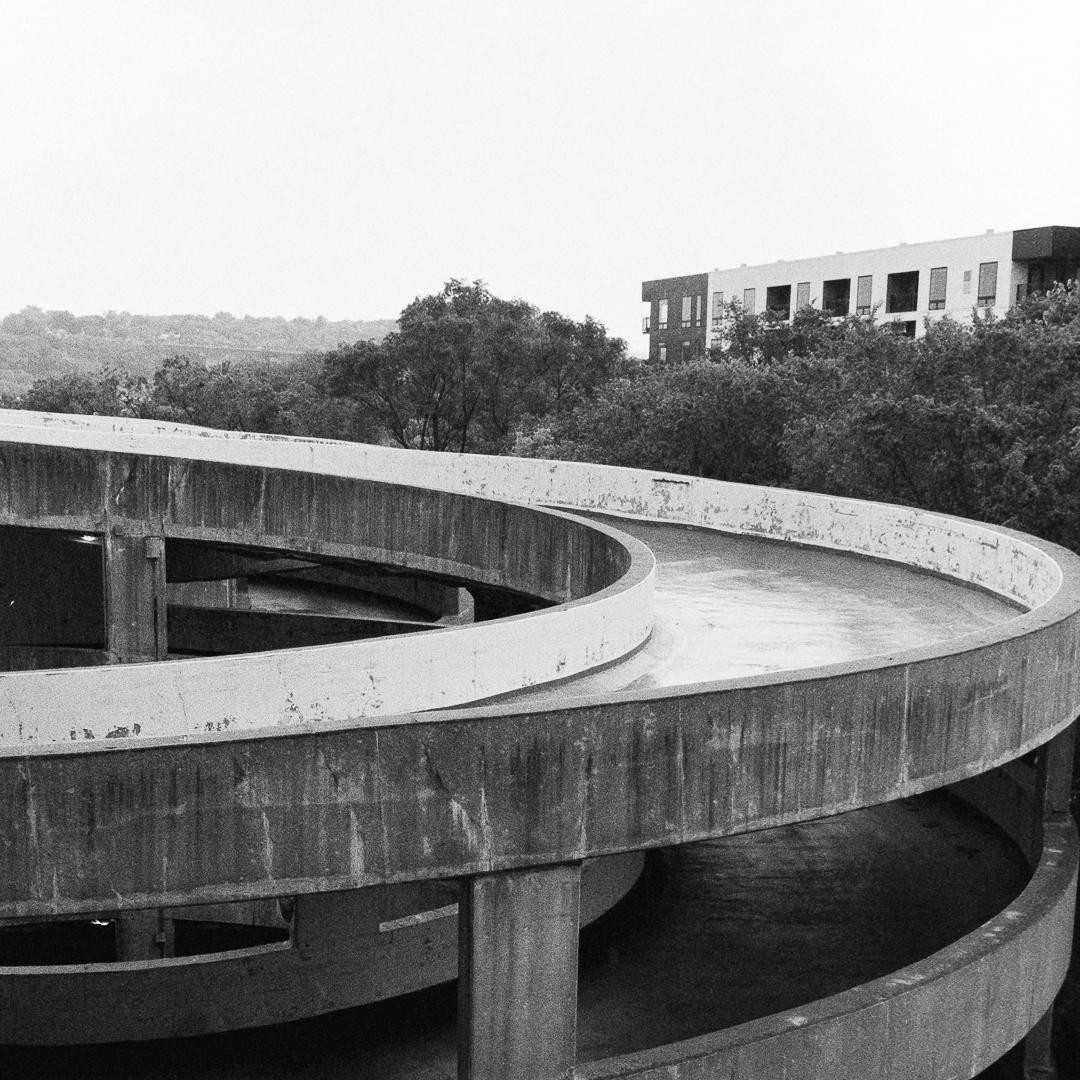 A concrete parking garage ramp curves upwards with trees and a building in the background.