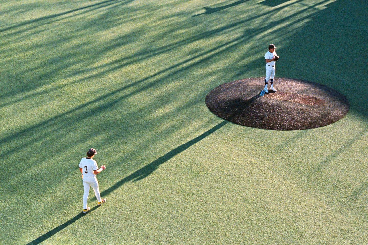 Two baseball players stand on a green field with long shadows cast across it.