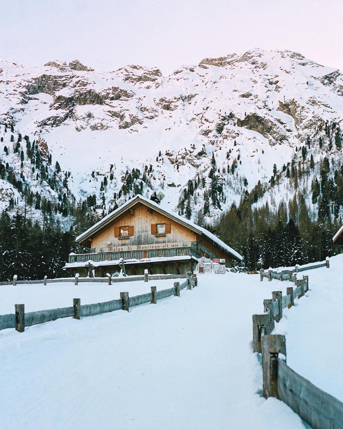 A wooden cabin with a fence in front of it is surrounded by snow-covered mountains and pine trees.