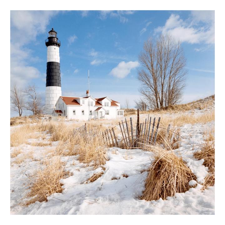 A black and white lighthouse stands next to a white house with a red roof on a snowy hill with dry grass and a wooden fence.