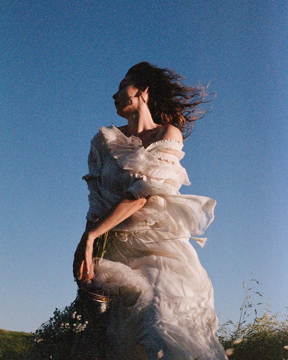 A woman in a white dress stands in a field with her hair blowing in the wind under a clear blue sky.