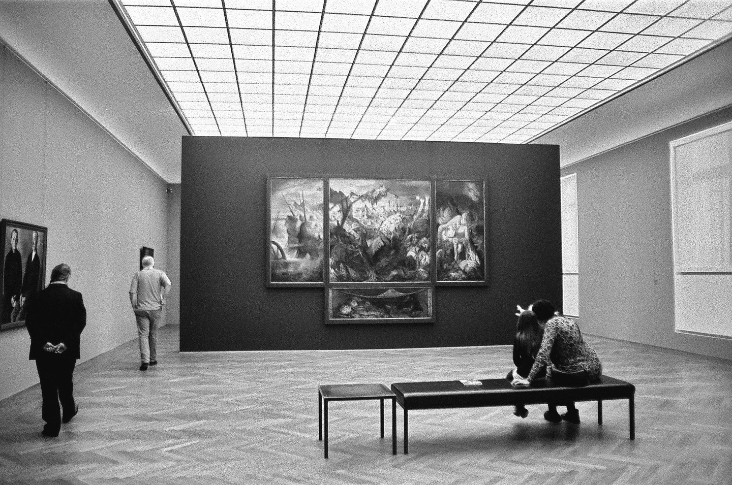 Two people sit on a bench in a museum gallery while two men walk past a large painting.