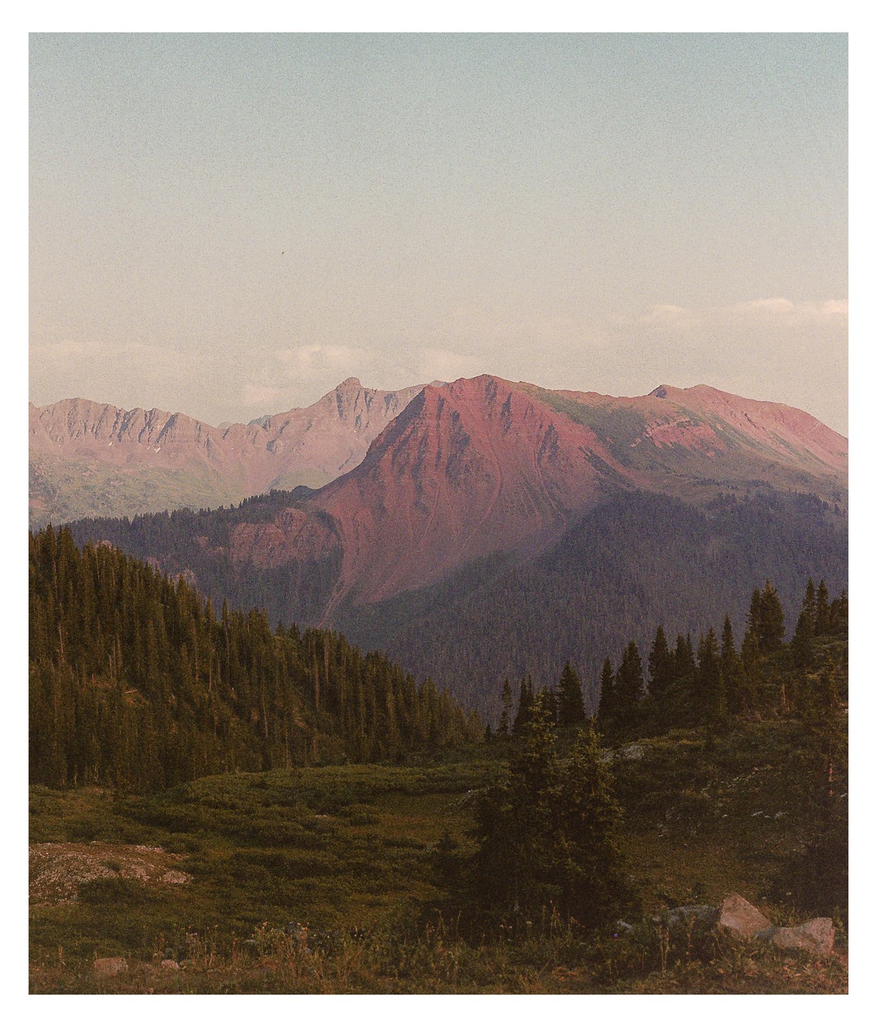 A vast mountain landscape features a prominent red mountain peak surrounded by green forests under a pale sky.