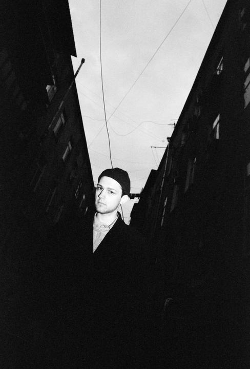A man wearing a beanie and coat stands in a narrow alleyway between two buildings with overhead wires.