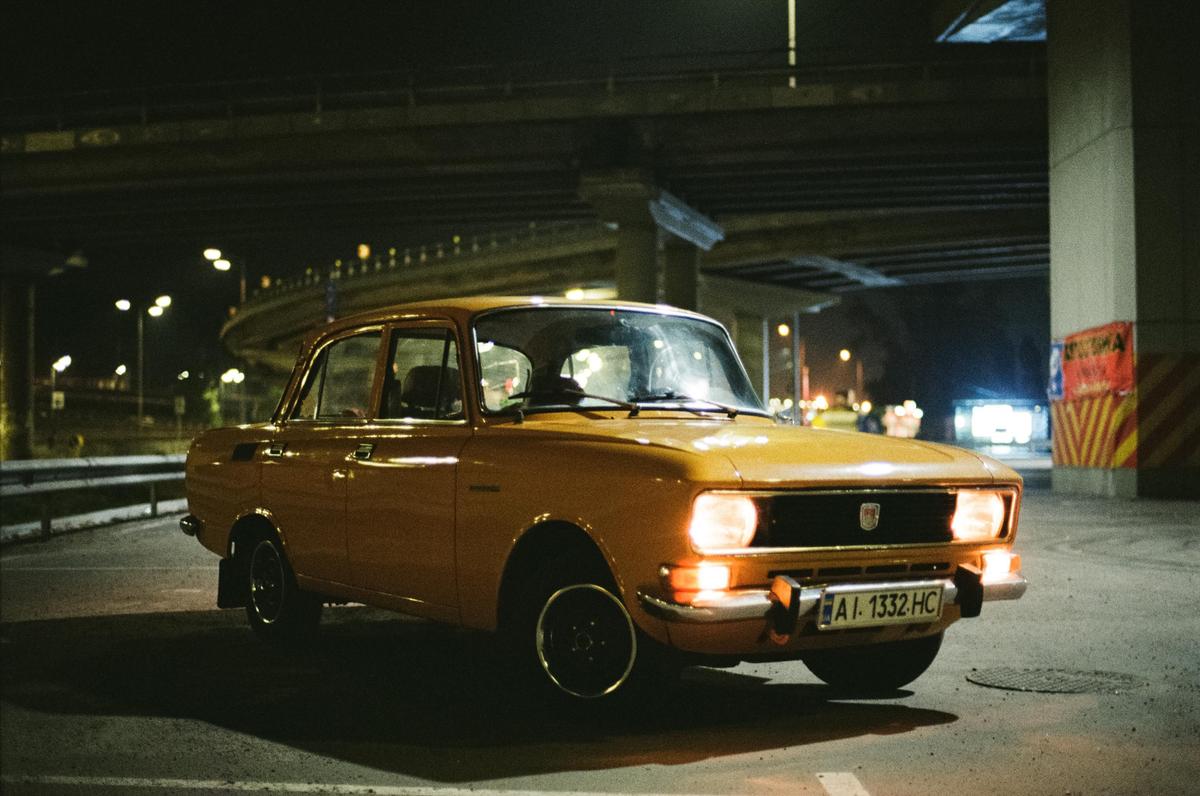 A yellow car with its headlights on is parked under an overpass at night.