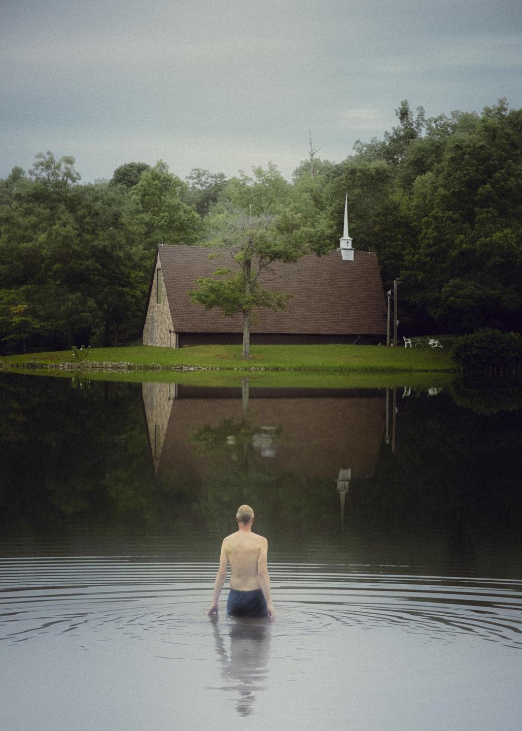 A man stands in a body of water with a church and trees reflected in the water.