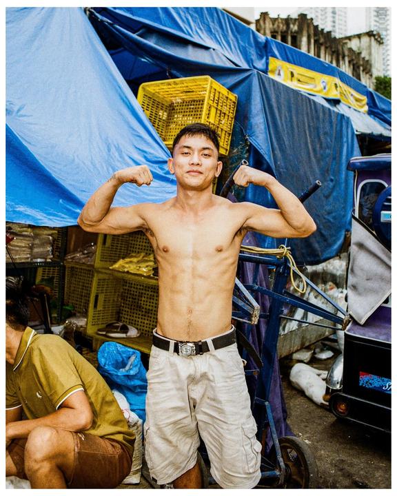 A man flexes his biceps while standing shirtless in front of blue tarps and yellow crates.