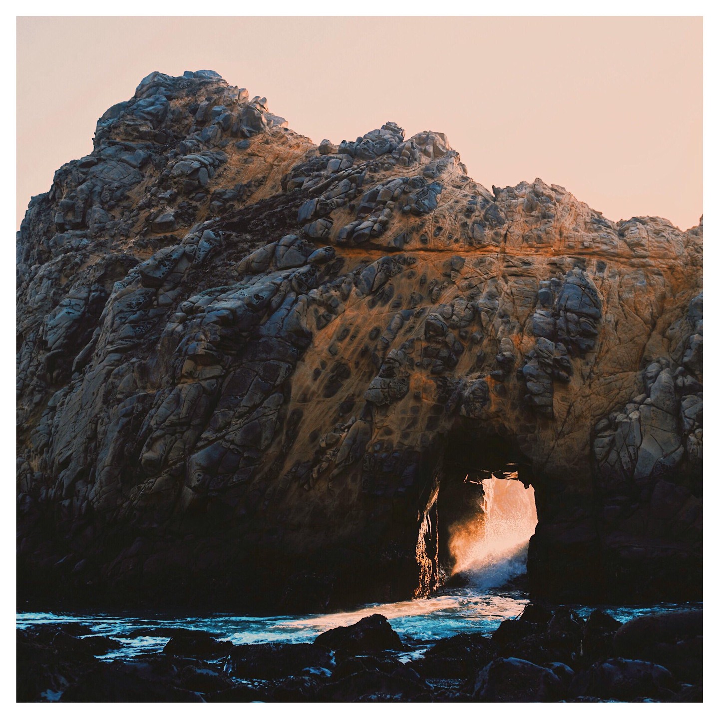 Sunlight shines through a natural arch in a large rock formation by the ocean.