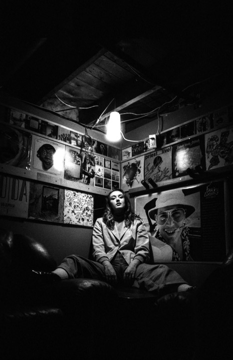 A woman sits on a couch in a dimly lit room adorned with album covers and posters.