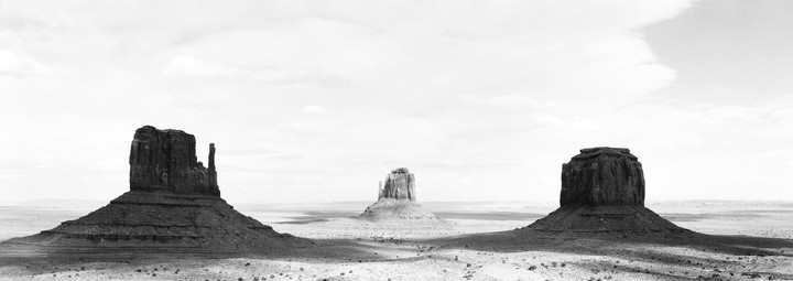 Three large rock formations stand in a desert landscape under a cloudy sky.
