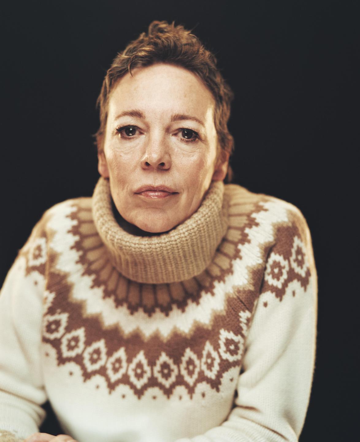 A woman with short hair wears a patterned turtleneck sweater and looks directly at the viewer.