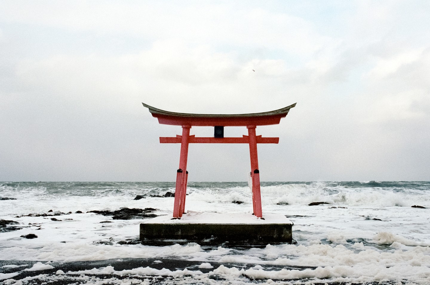 A red torii gate stands in the ocean with waves crashing around it and snow on the ground.