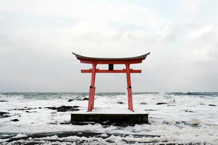 A red torii gate stands in the ocean with waves crashing around it and snow on the ground.