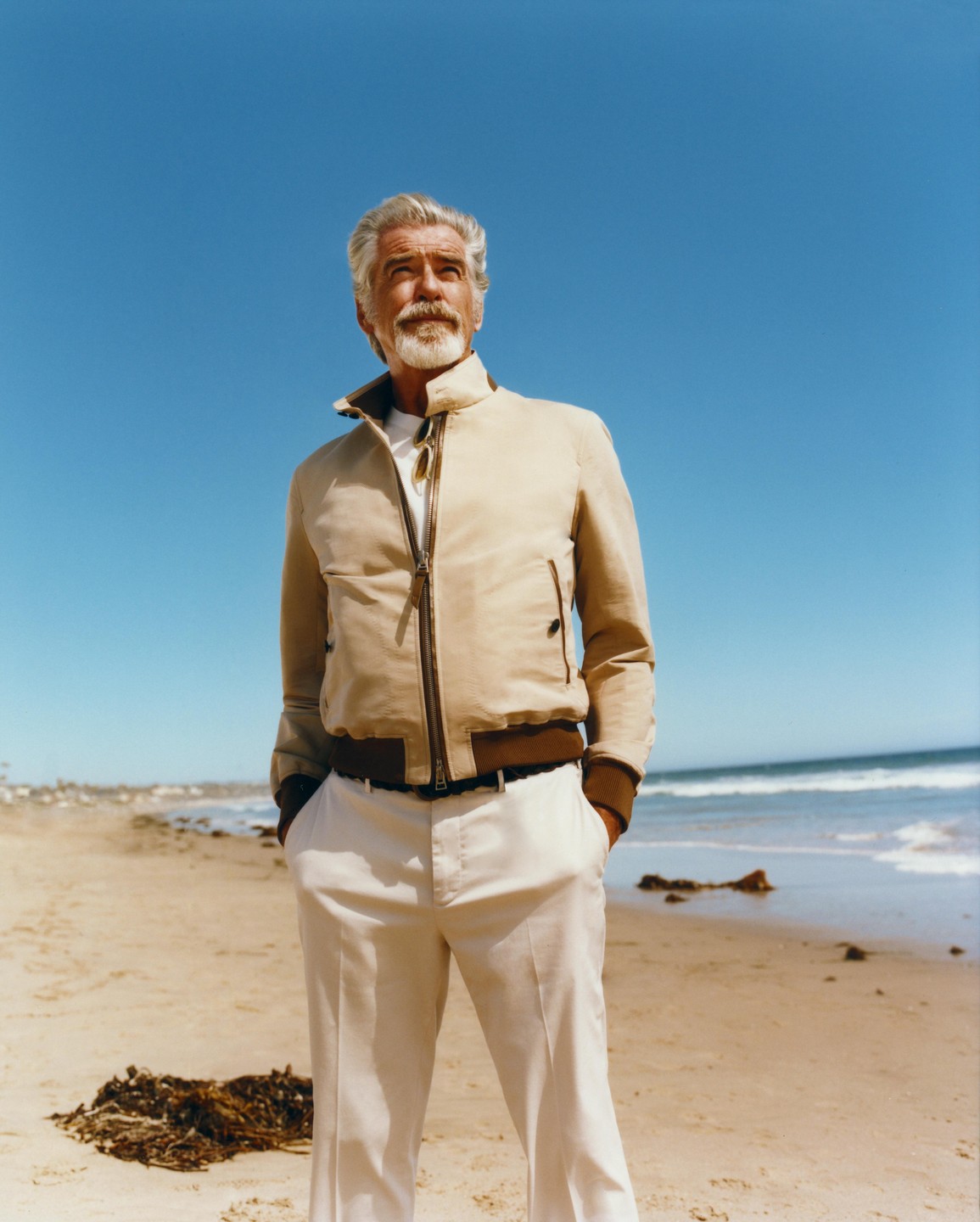 A man with a beard and gray hair stands on a beach looking up at the sky.