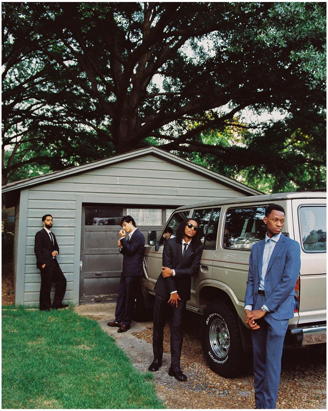 Four men in suits stand around a vehicle next to a garage with a large tree overhead.