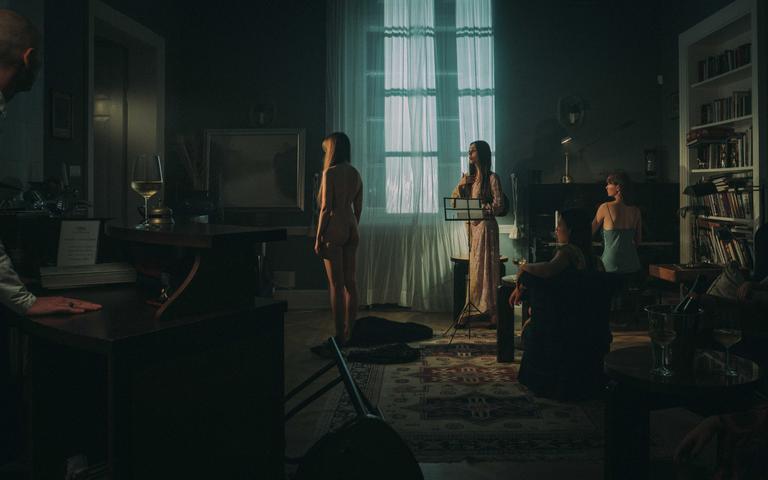A group of people are gathered in a dimly lit room with a large window and a bookshelf.