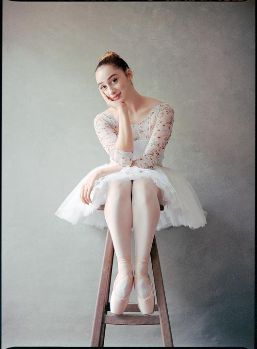 A ballerina in a white tutu and floral leotard sits on a wooden stool, resting her chin on her hand.
