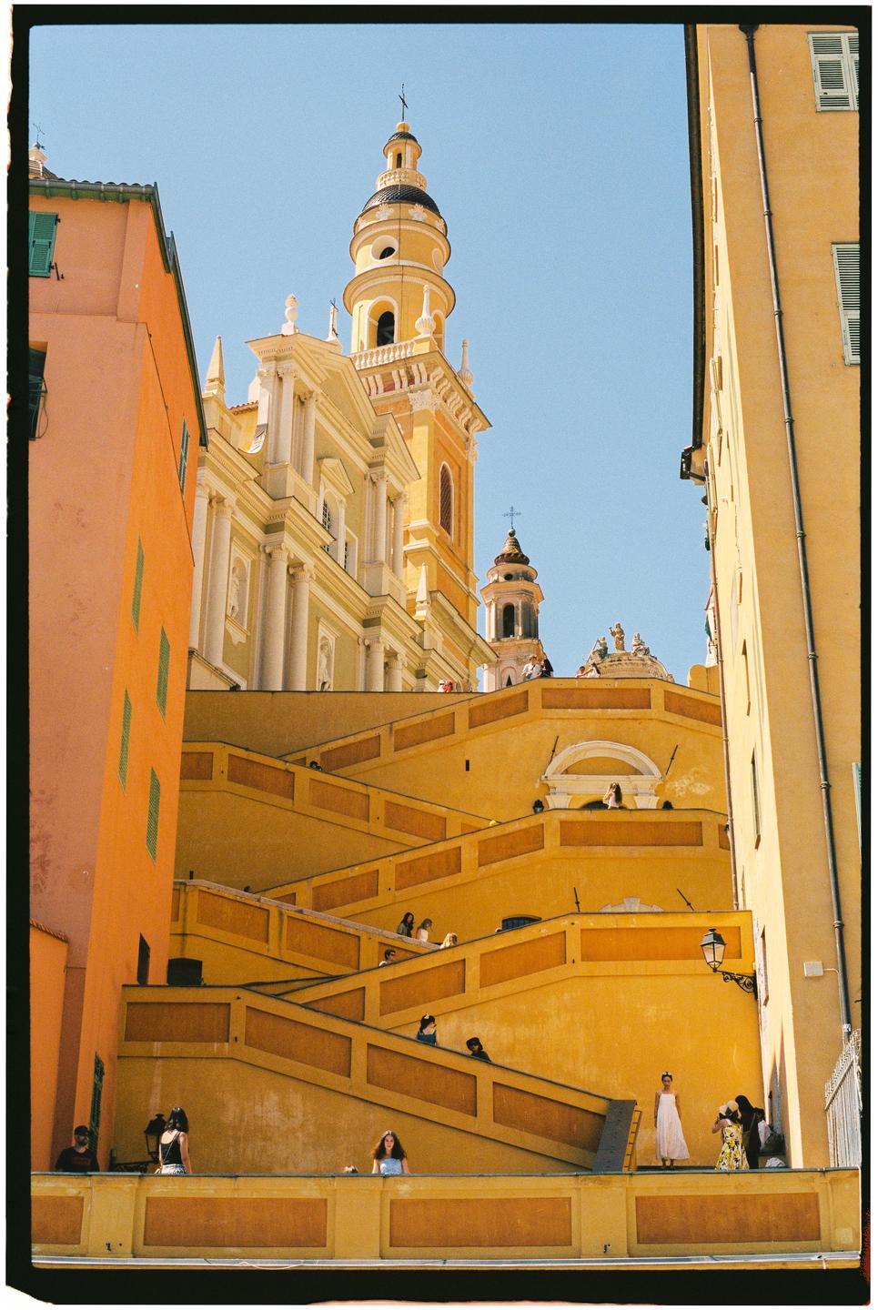 A tall yellow church tower rises above a complex system of yellow staircases and buildings.