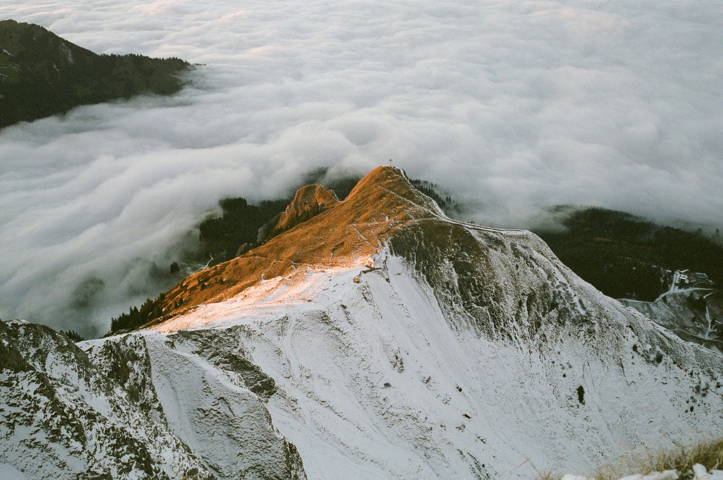 A snow-covered mountain peak rises above a sea of clouds, with a cross visible at its summit.
