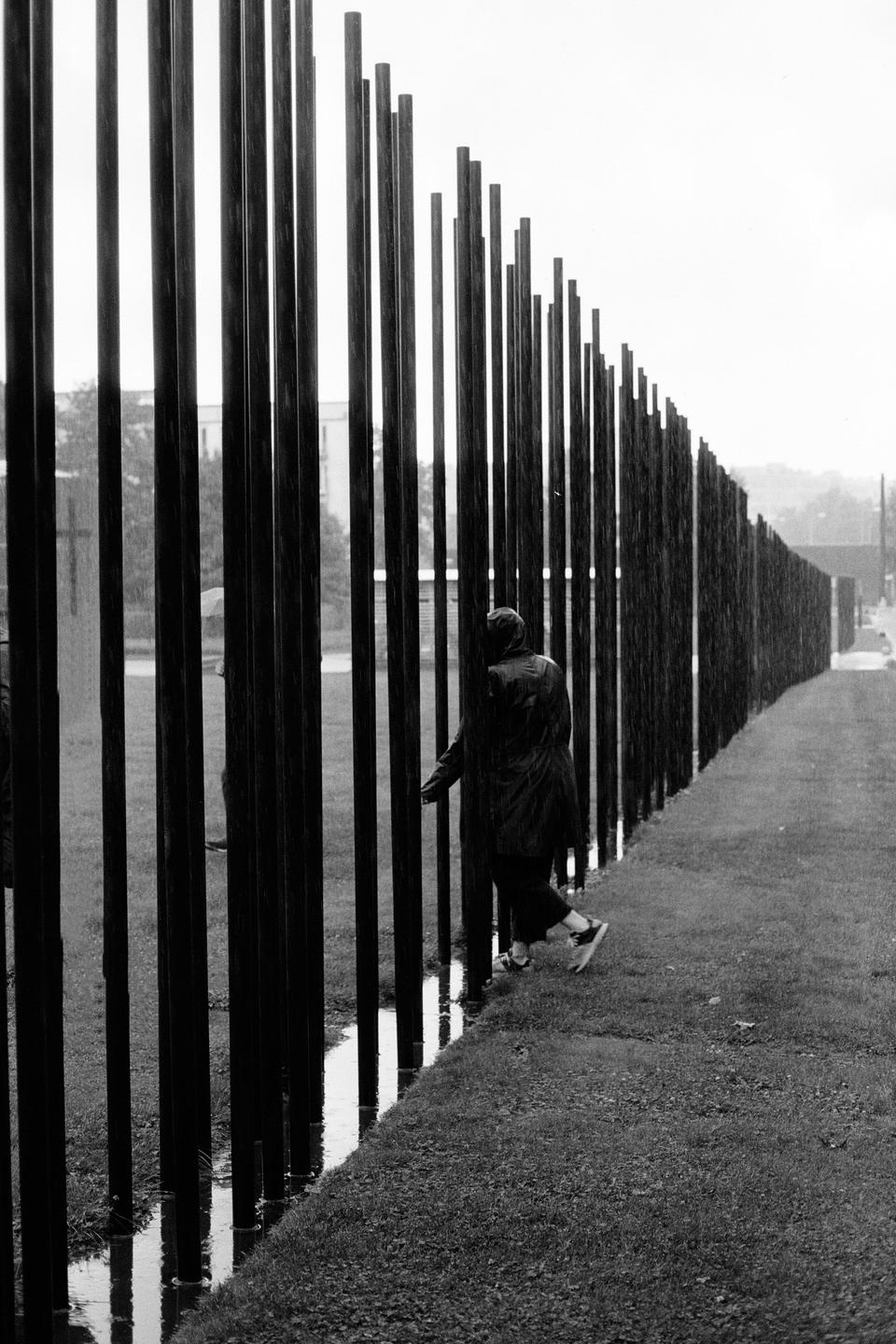 A person in a raincoat walks along a fence made of vertical metal bars on a rainy day.