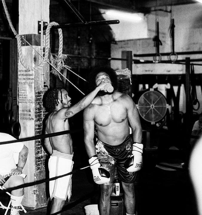 A man in boxing gloves drinks from a bottle while another man holds it for him in a boxing ring.
