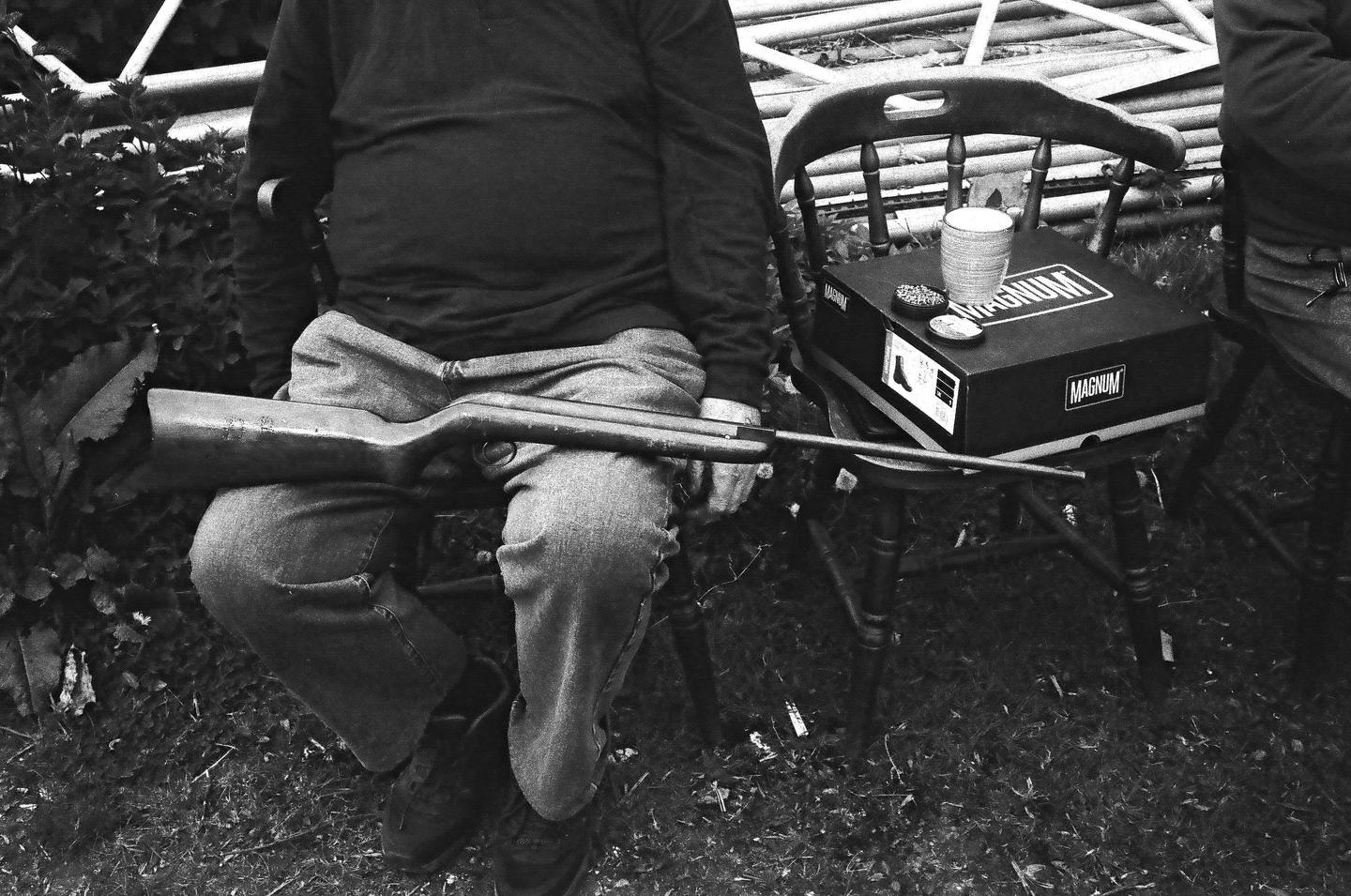 A man sits outdoors with a rifle across his lap and a box on a chair next to him.