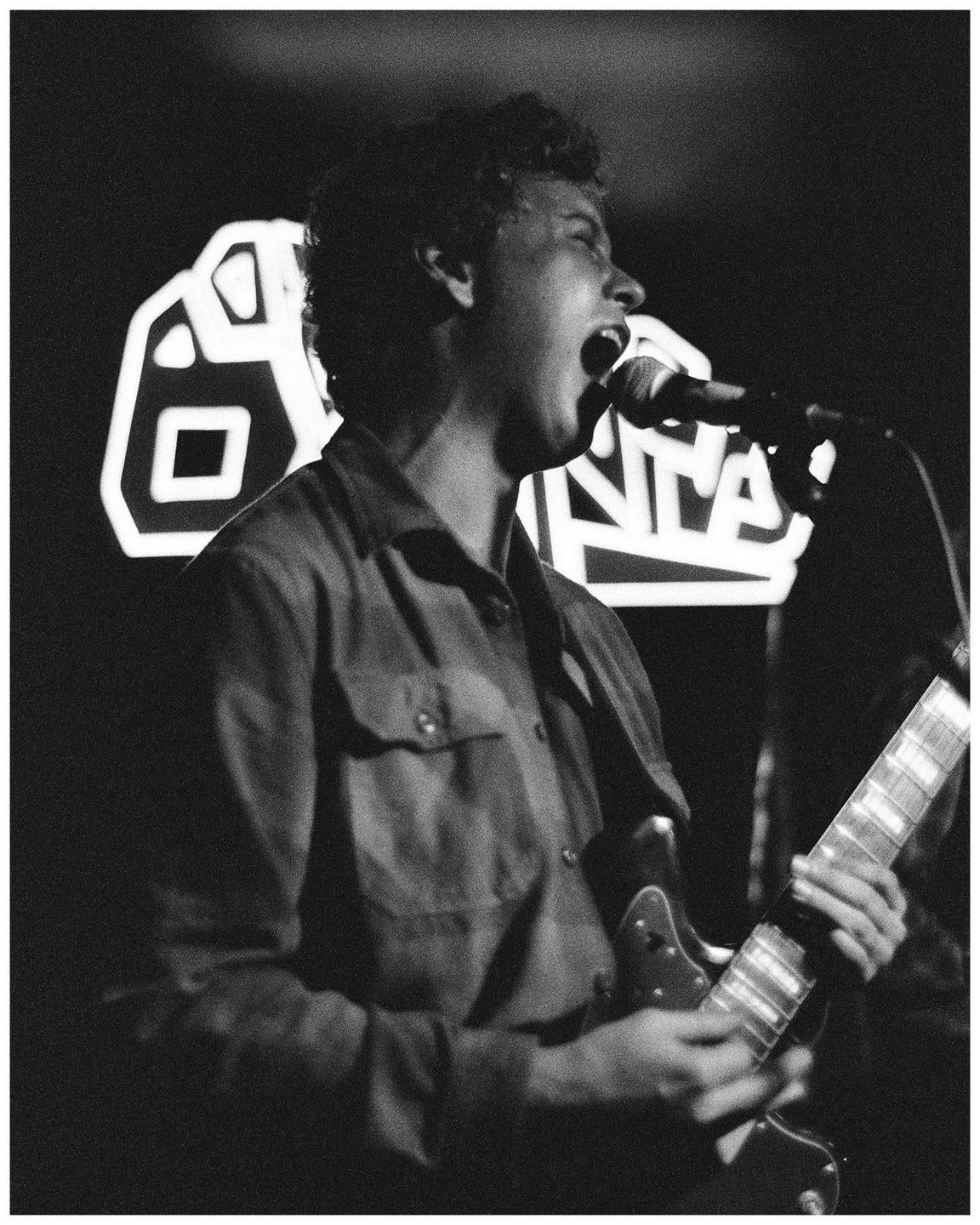 A man sings into a microphone while playing a guitar on a stage with a neon sign in the background.