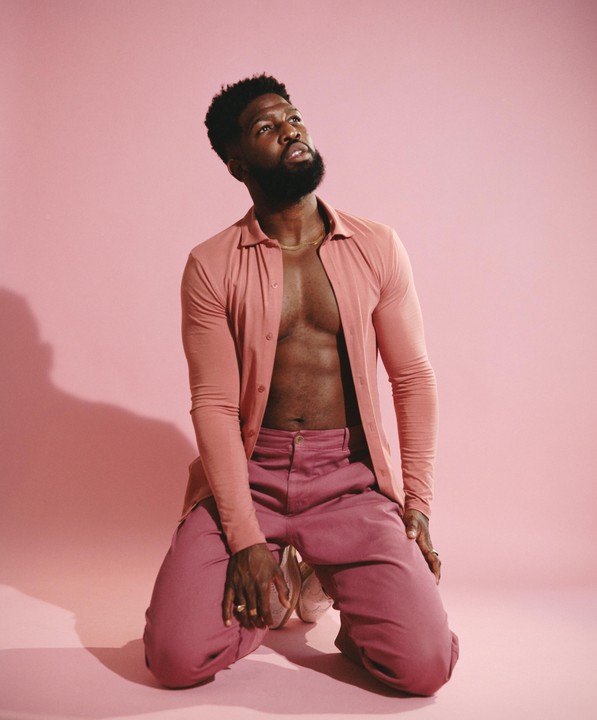 A man kneels on a pink surface, looking up, wearing a pink shirt and pants.