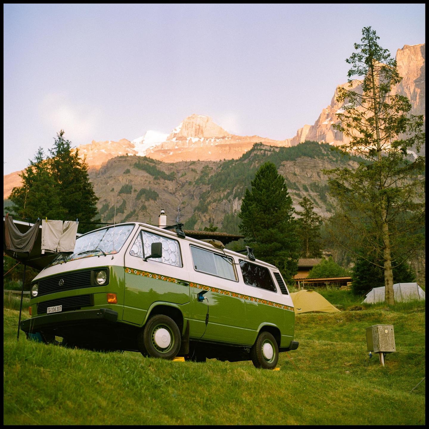 A green and white camper van is parked on a grassy hill with mountains in the background during sunset.