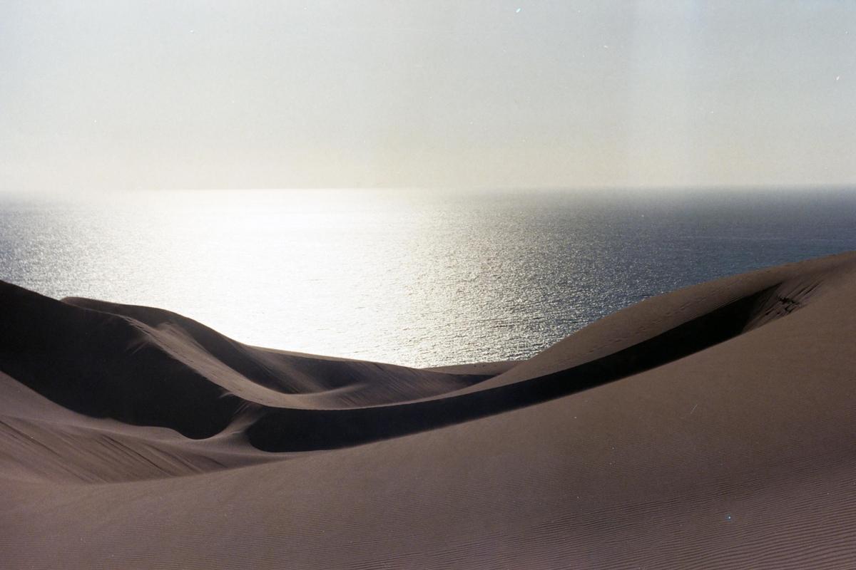 Sand dunes rise from the foreground with a body of water in the background reflecting the bright sky.